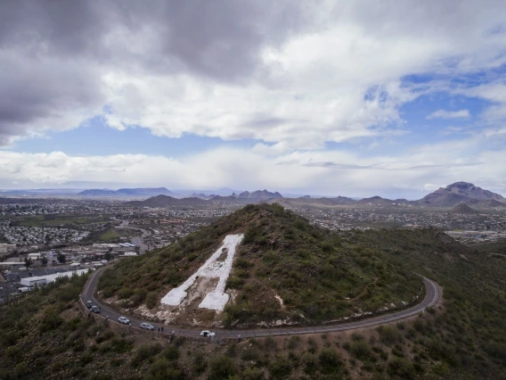 This image depicts a hillside with a large white letter "A" painted on it, surrounded by a winding road with several cars parked along the way. In the background, there is a vast cityscape extending into rolling desert mountains, under a partly cloudy sky.