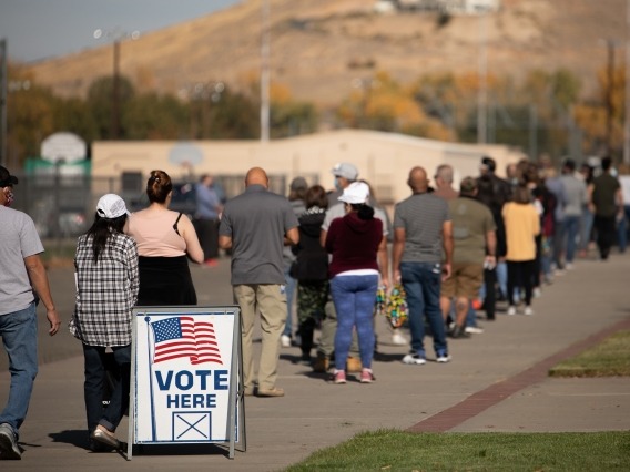A line of people facing away are standing on concrete in front of a school. There is a white sign with an American flag that says &quot;Vote Here&quot;