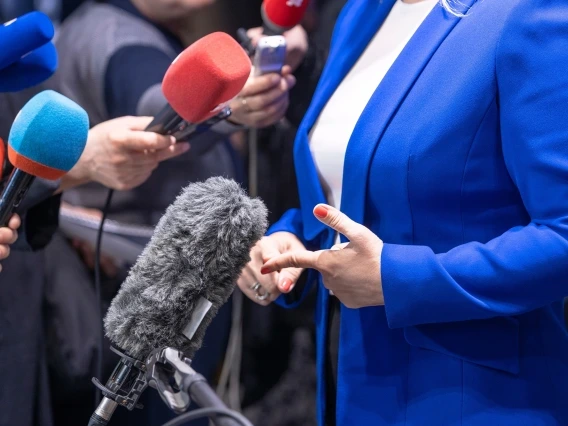 A close-up image of a person in a blue suit jacket being interviewed by the media. Several colorful microphones, including red and blue, are positioned toward the individual, who is gesturing with their hands while speaking. The focus is on the microphones and the person's torso, with the faces of reporters blurred in the background.