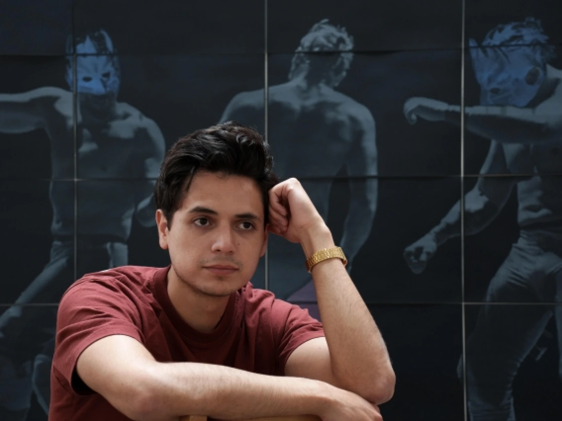 A man sits in front of a blue backdrop on which pictures of luche libre wrestlers are printed.