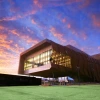 A modern, angular building with a geometric design situated against a dramatic sky during sunset. The structure features large glass windows framed by brown, asymmetric panels. The upper section of the building extends outward, creating an overhang supported by columns. The sky behind the building is a vibrant mix of orange and purple hues, with scattered clouds reflecting the sunset colors.
