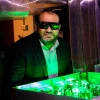 A man wearing safety glasses and a suit observes a green-lit optical experiment with lasers and mirrors in a laboratory setting.