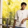 A young man with short dark hair leans casually against a railing, smiling warmly. He wears a white T-shirt, silver wristwatch, and a pendant necklace, with vibrant yellow trees and natural light in the background.